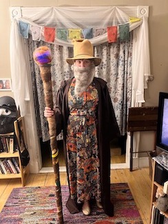 Rebecca dressed as wizard with a long grey beard, pointy hat, colorful robe, brown cloak, and tall staff topped with a blue orb.